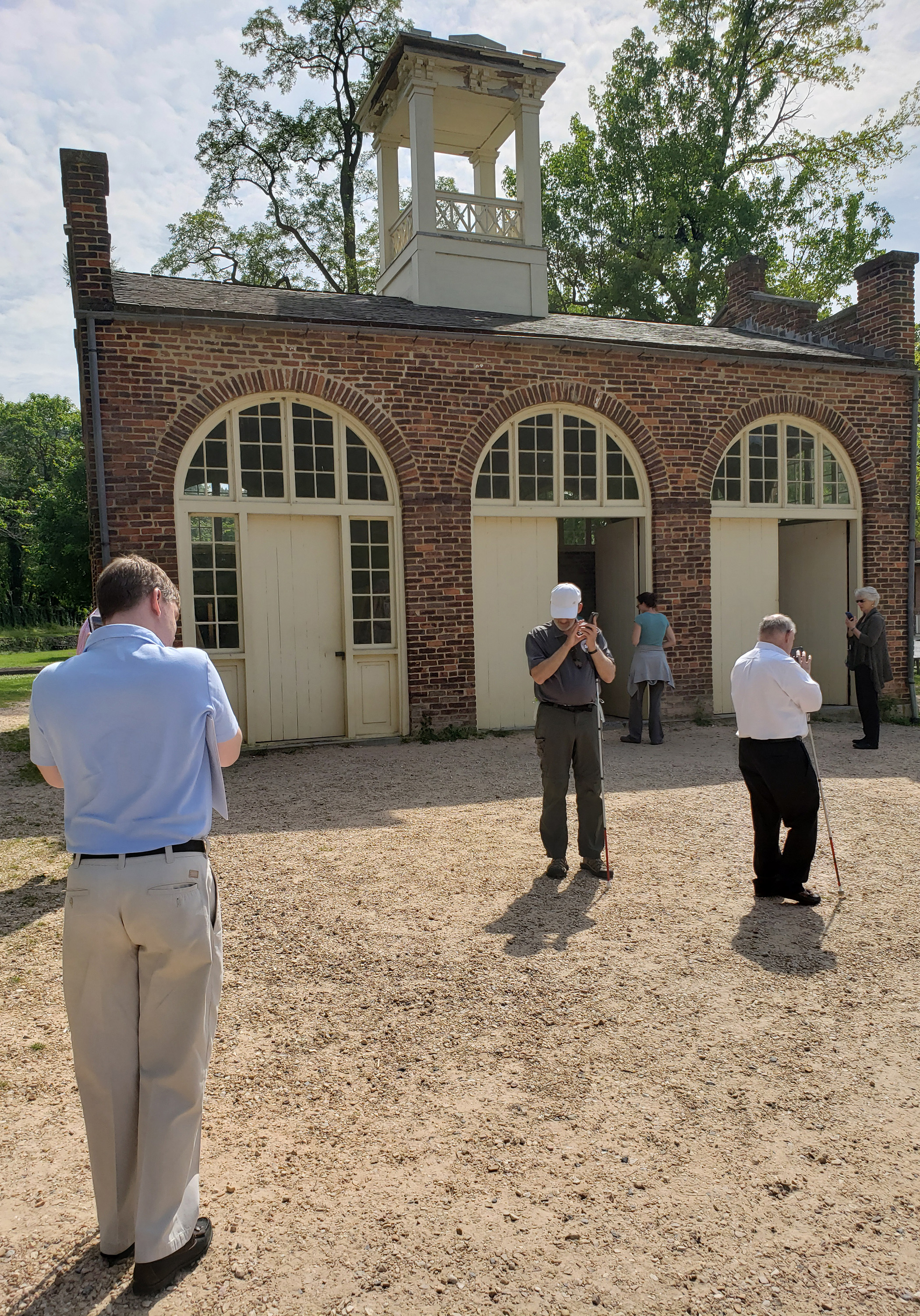 DESCRIBING: A vertical color photograph. DESCRIPTION: From left to right in the foreground, Eric Bridges, American Council of the Blind's Executive Director, Doug Powell, a member of ACB’s Rehabilitation Task Force, and Pat Sheehan, Director of the 508 Program Office at the U.S. Department of Veterans Affairs, use the UniD app to explore John Brown's Fort with U.S. National Park Service staff members at Harpers Ferry National Historical Park in West Virginia. In this image, the three ACB members are listening to their smartphones on the dirt patch outside of the fort. The fort – a one-room, single-story brick building – has three identical entry doors, each surrounded by an arch of window panes. Two of those doors are propped open for visitors to go in and out.