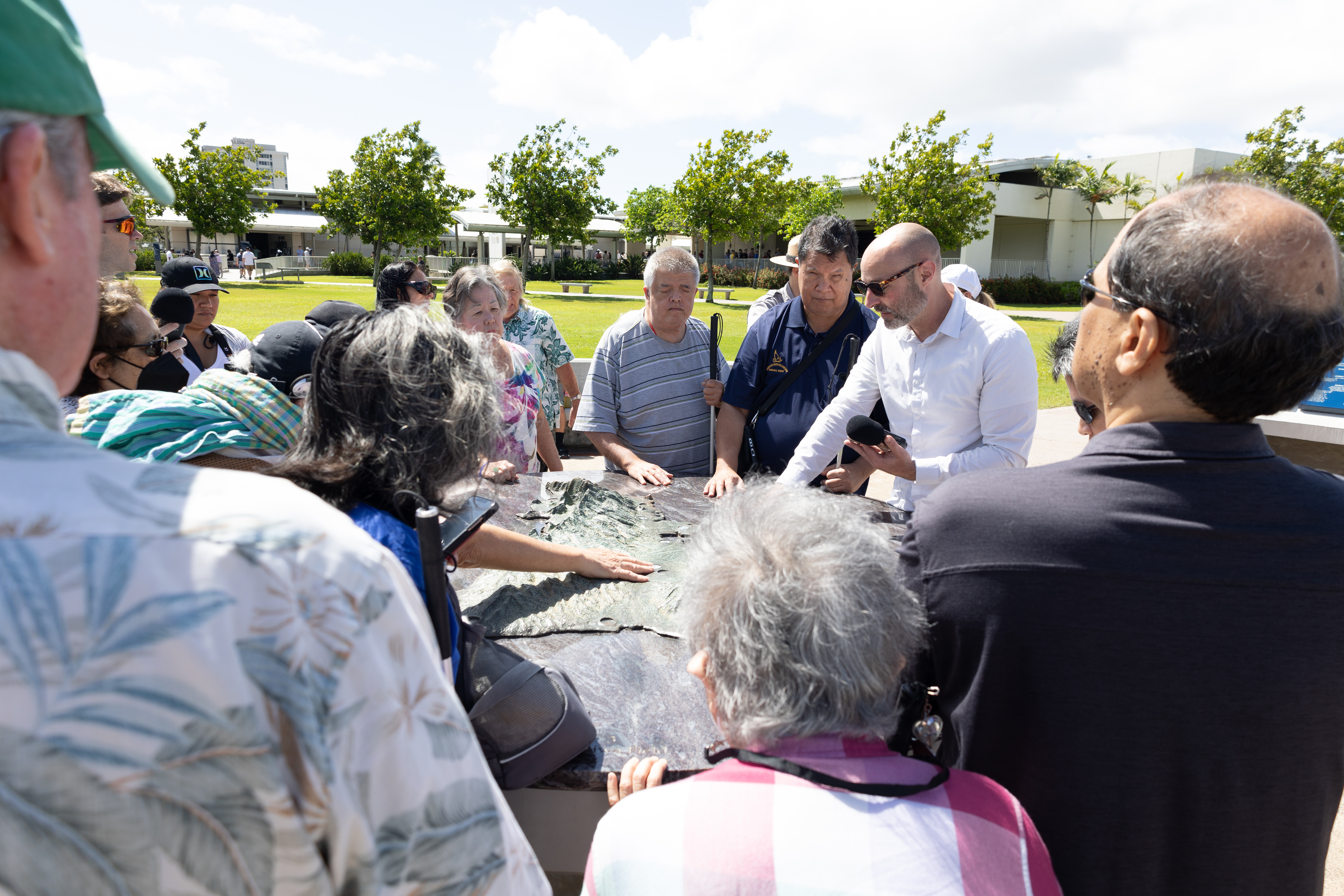 DESCRIBING: A horizontal color photograph. DESCRIPTION: Dr. Jordan Frith from Clemson University joined the research team in Honolulu to help conduct field research at Pearl Harbor National Memorial. In this horizontal color photograph, Dr. Frith is slightly to the center of the image, describing to a group of 15 research participants — as well as friends and family members serving as chaperones — how a Near-Field Communication Tag (NFC) works. For this experiment, our team placed a quarter-sized NFC tag on this map that could be accessed and heard via smartphones using the UniD mobile app. To use the tag, the listener just had to open the app, find the tag on the map, and place the phone on the tag. In this image, Dr. Frith is wearing a white collared shirt and sunglasses, and he is standing with the huge circular metal map about at his hip level. He is touching the map with his right hand and holding an audio-recording device in his left hand. Hawaii Association of the Blind members ring the table and are listening to the instruction, with more Pearl Harbor buildings and facilities, lined with trees, in the background.