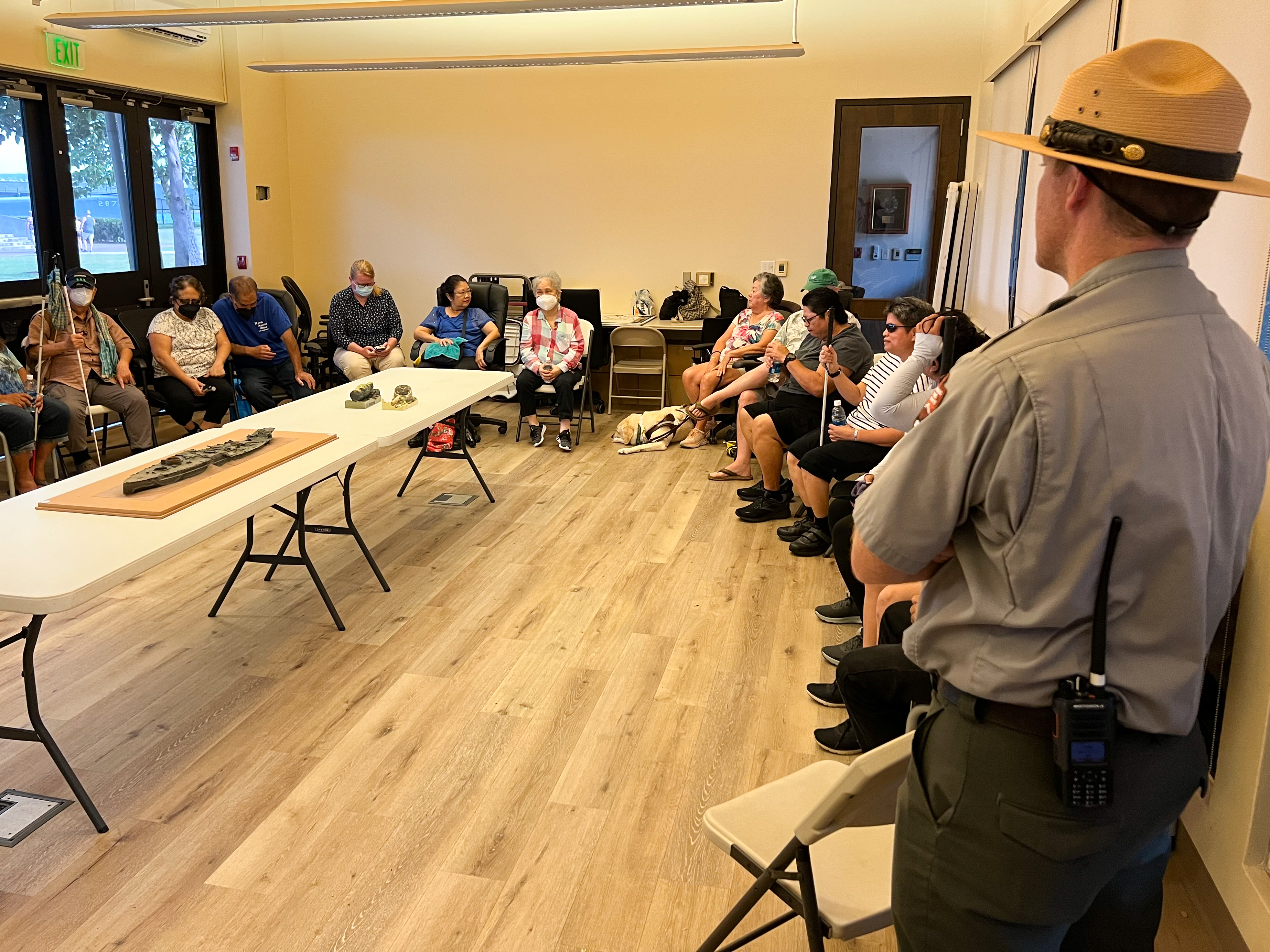David Kilton, Chief of Interpretation at Pearl Harbor National Memorial, is shown in the foreground, with his back to the camera, listening to a focus group discussion about Audio Description at the National Park Service site. About 20 Hawaii Association of the Blind members are sitting in a U-shaped formation, facing Kilton, engaged in the discussion, although not all of them are included in this particular image. Kilton is dressed in a formal National Park Service uniform, including a tan and round-brimmed hat and khakis, with his buttoned-up shirt a slightly paler color than the dark-green pants heʻs wearing. A long rectangular table in the middle of the room, in the middle of the U-shape made by the chairs, displays three tactile models that are being discussed during this session. Those are of a ship, a soda bottle, and a cooking pot. The HAB members are dressed in casual clothes, with some of them holding white canes and with one guide dog in the image, who is on the floor next to his companion.