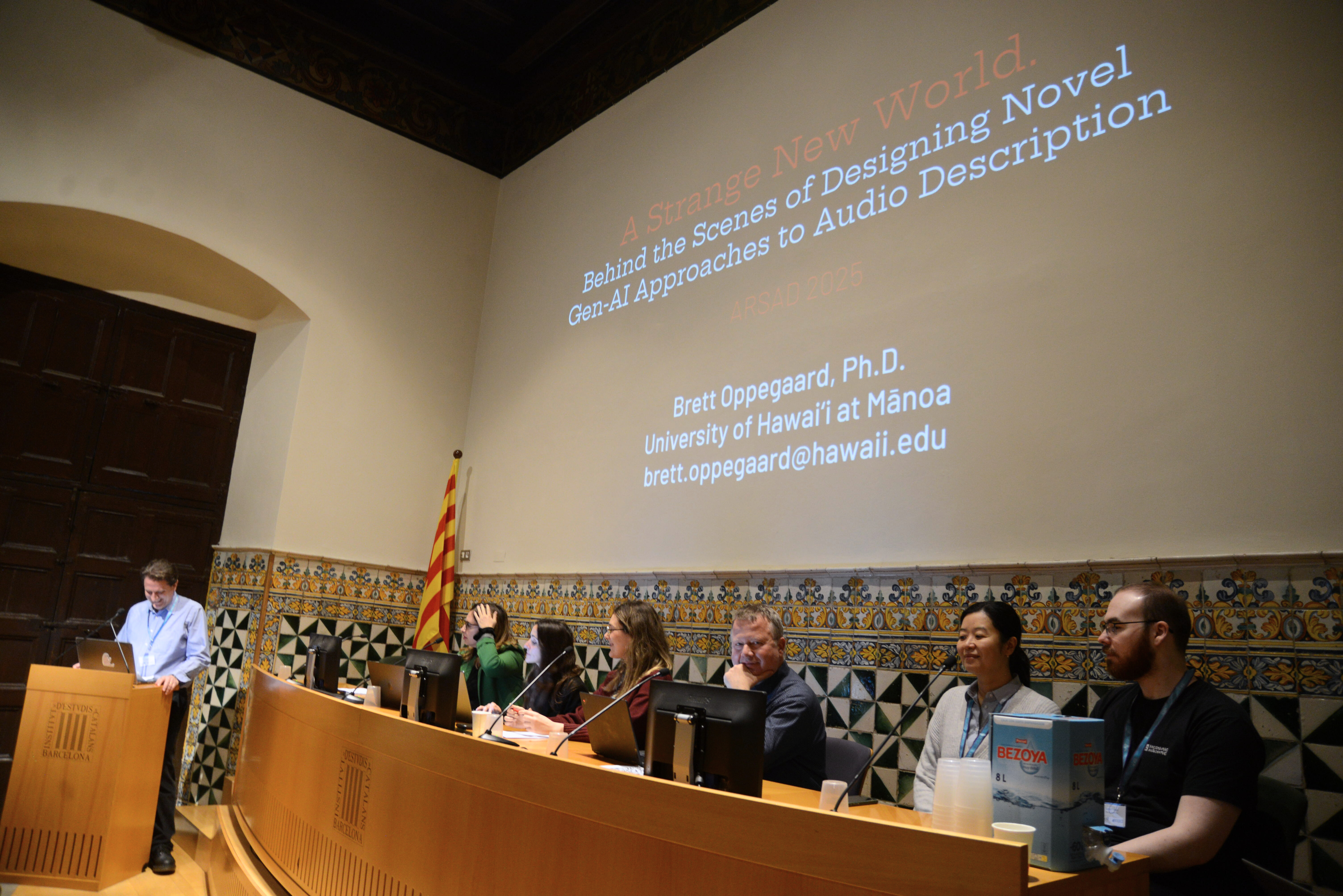 DESCRIBING: A horizontal color photograph. DESCRIPTION: Dr. Brett Oppegaard stands at a lectern, giving a presentation about Audio Description, in front of a panel of other researchers at the Advanced Research Seminar on Audio Description (ARSAD) conference in Barcelona, Spain. A large screen in the background states the name of his presentation: 'A Strange New World: Behind the Scenes of Designing Novel GenAI Approaches to Audio Description.'