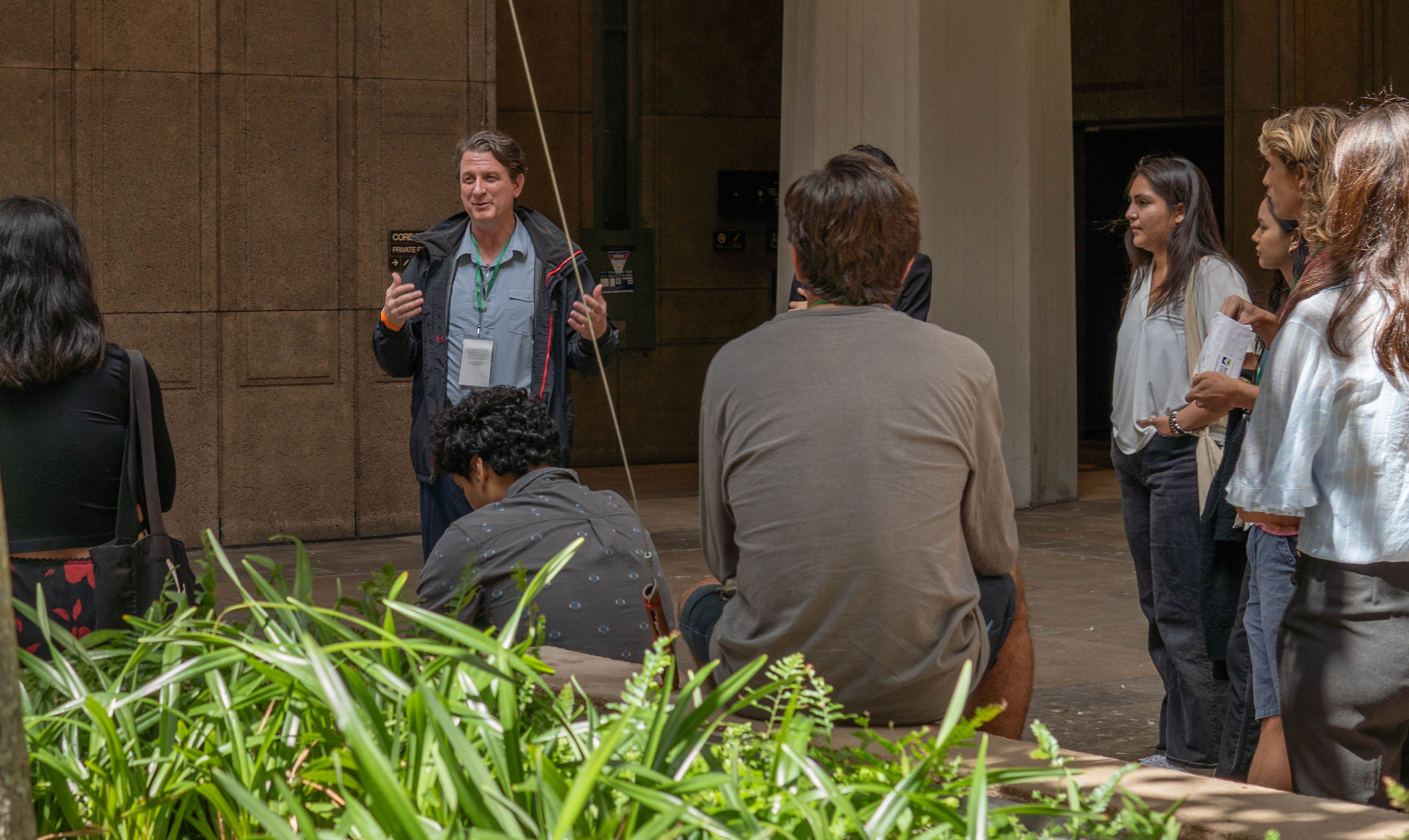 DESCRIBING: A horizontal color photograph. DESCRIPTION: Dr. Brett Oppegaard is standing among a crowd of students, talking to them about how to be better writers. He is wearing a black rain jacket, light-blue collared shirt, and a lanyard with his identification. The scene is at the Hawai‘i state capitol building, inside the rotunda, which is a large open area. The only visible parts of the environment that can be seen arre a large stone pillar and a stone wall. There are about a half-dozen students around Dr. Oppegaard, mostly with their backs to the camera, facing their teacher.