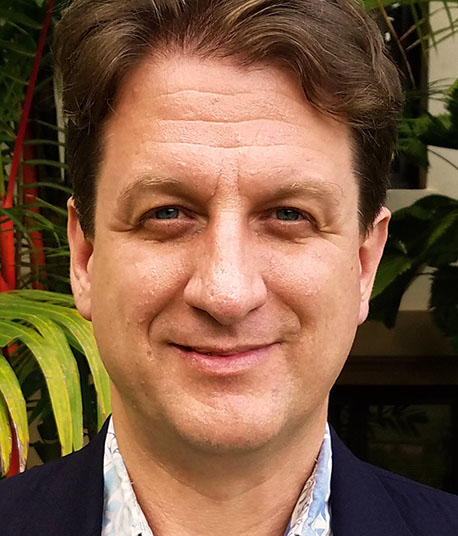 In this small vertical portrait, I am shown from my shoulders up to roughly the middle of my forehead. I am a 54-year-old American man, white in the abstract but with ancestral origins in the United Kingdom and Norway. Indicating those roots visually, I have blue eyes, a pinkish-tan skin with a red undertone, and dark brown hair. My hair is short on the sides but kept long on the top, showing some waviness to it. I do not have any facial hair beyond my eyebrows. The collar of my light-blue and patterned Aloha shirt shows just above my dark-blue suit jacket. On the UH campus, Aloha shirts are common, and almost no one wears a tie. So I’m dressed for that work environment. I am looking directly into the camera and expressing a subtle smile, with my lips slightly upturned at the corners. I am hoping to convey warmth and friendliness while still maintaining an authentic facial expression.