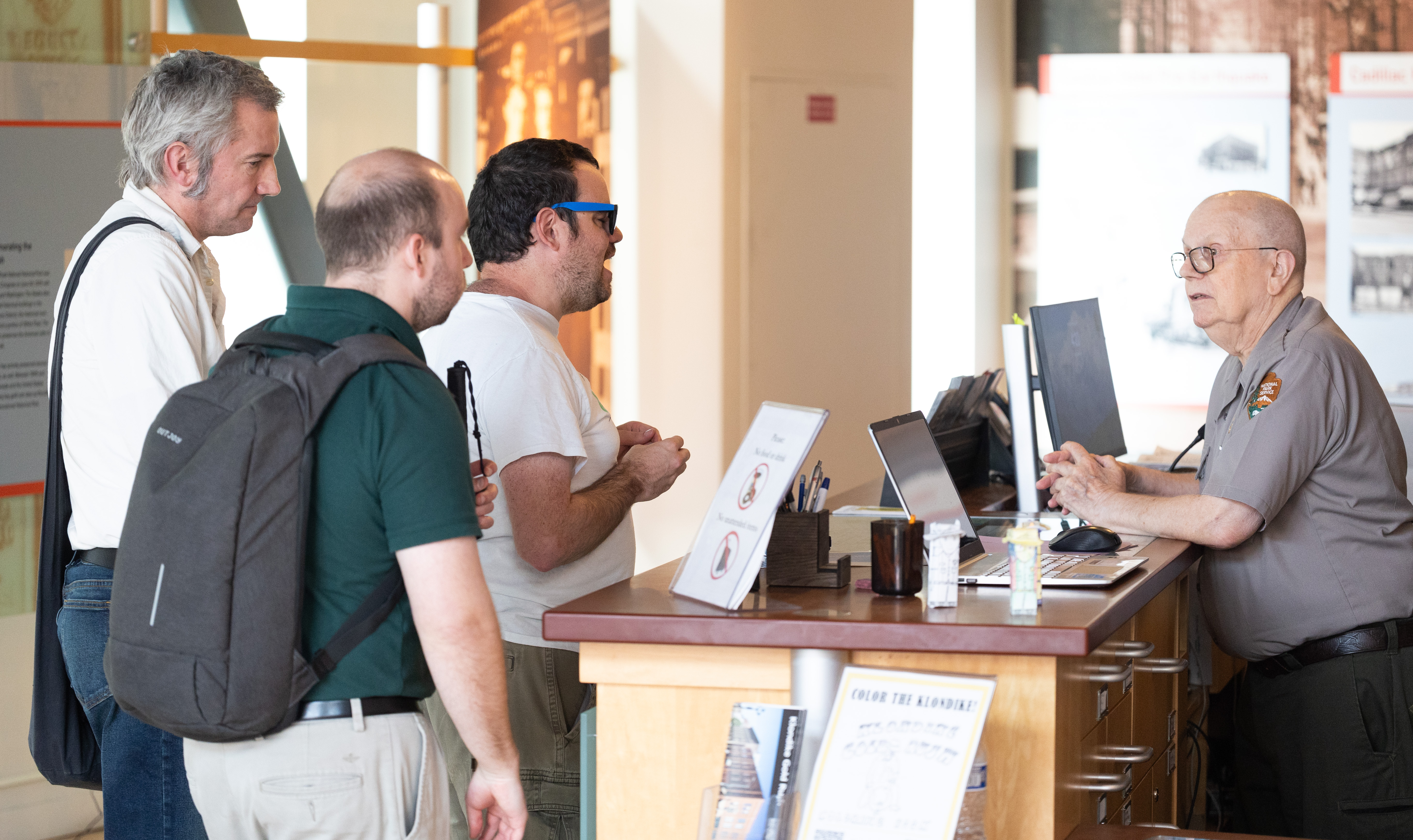 DESCRIBING: A horizontal color photograph. SYNOPSIS: Inside a bright visitor center, three visitors stand at a wood-topped counter facing a uniformed staff member. The staffer, an older man in glasses and a gray short-sleeve shirt with an arrowhead-shaped shoulder patch, works behind two open laptops. The customers lean in and talk, one holding a cane upright. Posters and exhibit panels glow softly in the background, framing the courteous exchange. IN-DEPTH DESCRIPTION: Foreground, left to right: A gray-haired man with a shoulder bag stands slightly behind two others. Next, a man with a close-cropped head, green polo, and a large dark backpack faces the counter; his right hand rests near a slim cane with a black handle, held vertically. Closest to the counter, a man in a white T-shirt and bright blue-framed sunglasses gestures mid-conversation, his head turned toward the staffer. Right side: Behind the counter, an older man in glasses sits or leans forward, hands clasped near a keyboard. He wears a gray uniform shirt with nameplate and an arrowhead-shaped patch on the sleeve. Two laptops, a computer mouse, a pencil cup, and a small tent sign with circular icons sit along the counter’s edge. Background: Light-colored walls and exhibit panels with historic photos create a museumlike atmosphere. Warm daylight and soft focus keep attention on the people. The overall mood is helpful and attentive, as if the group is asking for help and support.