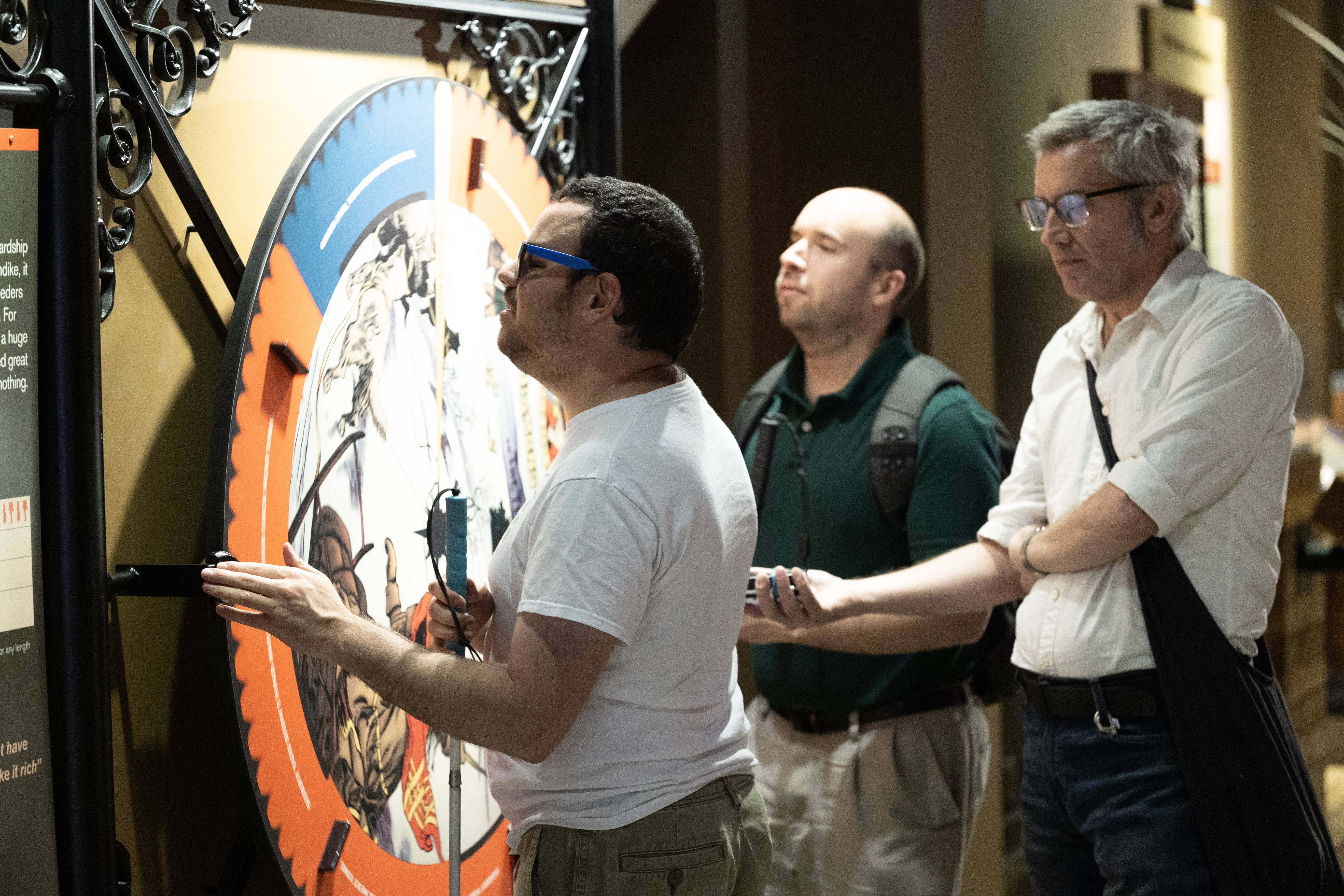 DESCRIBING: A horizontal color photograph. DESCRIPTION: Three men are standing next to a museum exhibit that includes a large wheel that can be spun to show the low odds of finding gold during the Klondike Gold Rush. The man on the left, Bob Cavanaugh, is touching the wheel with his left hand and feeling the part of the exhibit that points to the wheel. Cavanaugh is wearing a white t-shirt and sunglasses. He has short brown hair, white skin, and a bit of beard stubble. Timothy Breitenfeldt, center, is wearing a green collared shirt and khaki pants, with a black backpack. He is facing the wheel, too. He has white skin, short brown hair on the sides of his head, and bead stubble as well. Matt Bullen, right, is wearing a white-collared shirt, black pants, and has a bag around his shoulders, allowing him to carry his research equipment. He is holding an audio recorder in his right hand, extended toward Cavanaugh, capturing his comments. Bullen has short gray hair, white skin, and glasses.