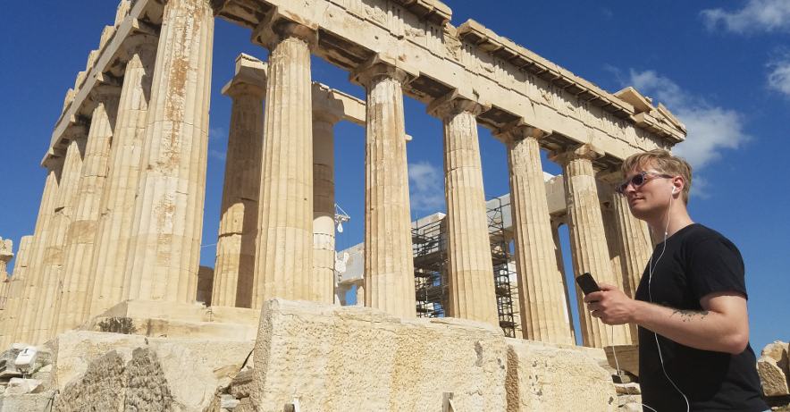 Access Hound CTO Joe Oppegaard, with smartphone in hand, at the base of the Acropolis in Athens, Greece, testing our Audio Description systems. Joe is wearing a black shirt and dark sunglasses, contrasting with his blond hair and white earbuds, which are connected to his smartphone, as he listens to content on the phone underneath the massive marble columns at the remains of the Parthenon. The Access Hound team has worked with the University of Piraeus in Athens to audio describe parts of the Acropolis as well as various other major public attractions around the world.