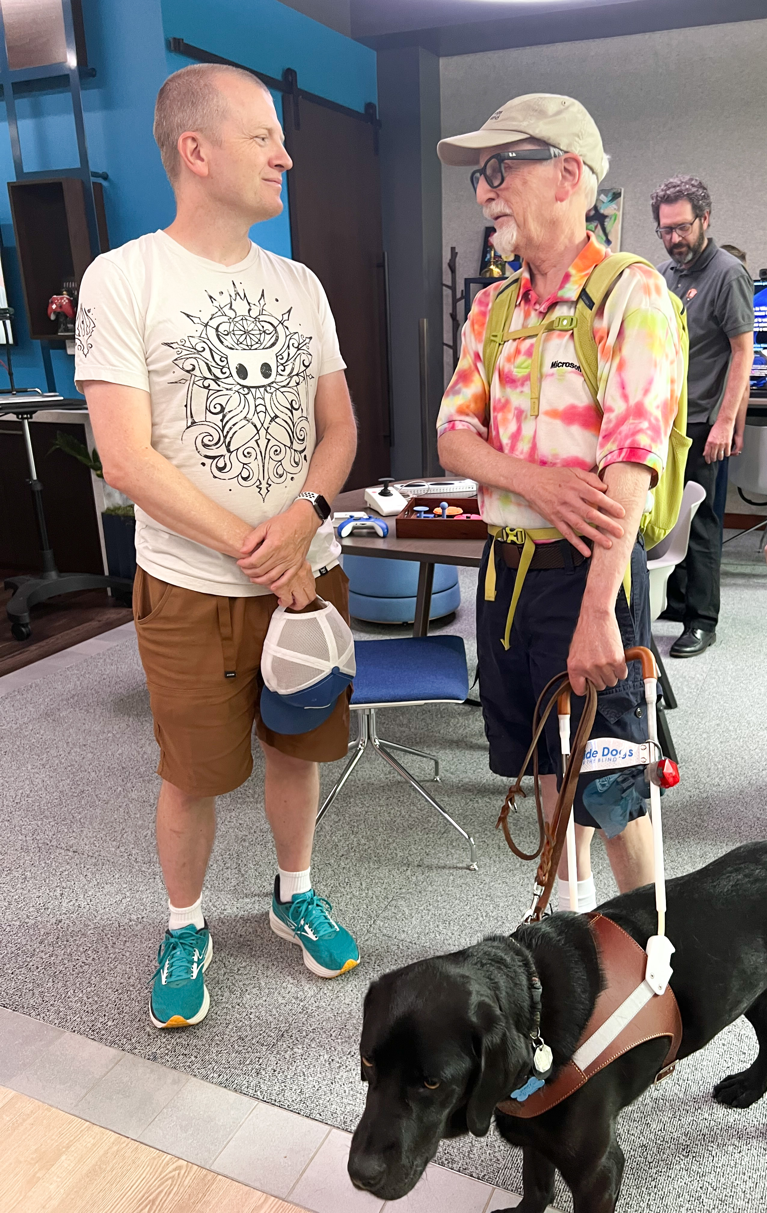 Stewart McCullough, left, one of the co-founders of Access Hound, stands in the Microsoft Inclusive Tech Lab facing collaborator Jeff Abrams, who is blind and accompanied by a guide dog. McCullough and Abrams are both white skinned and wearing a shorts and a t-shirt. McCullough's shirt is white, with tan shorts, and Abrams is wearing a tie-dyed shirt with black shorts. McCullough has a baseball hat in his hand, and Abrams is wearing one. Abrams also is wearing a pair of Meta Ray-Ban glasses, which have technology that helps him to gather information about the place he is in, like this lab. His dog is black and is wearing a harness with a rigid handle that Abrams is holding. In the background, there are various gadgets that were designed to make more-accessbile computing, such as adaptive joysticks and mice. Also in the background is Charles Beall, the superintendent of Klondike Gold Rush National Historical Park, who came to the lab with the Access Hound group. Beall is wearing a collared dark-green NPS shirt, with black pants. He is white, with short brown hair, glasses, and a full beard. He is looking over the gadgets on the table.