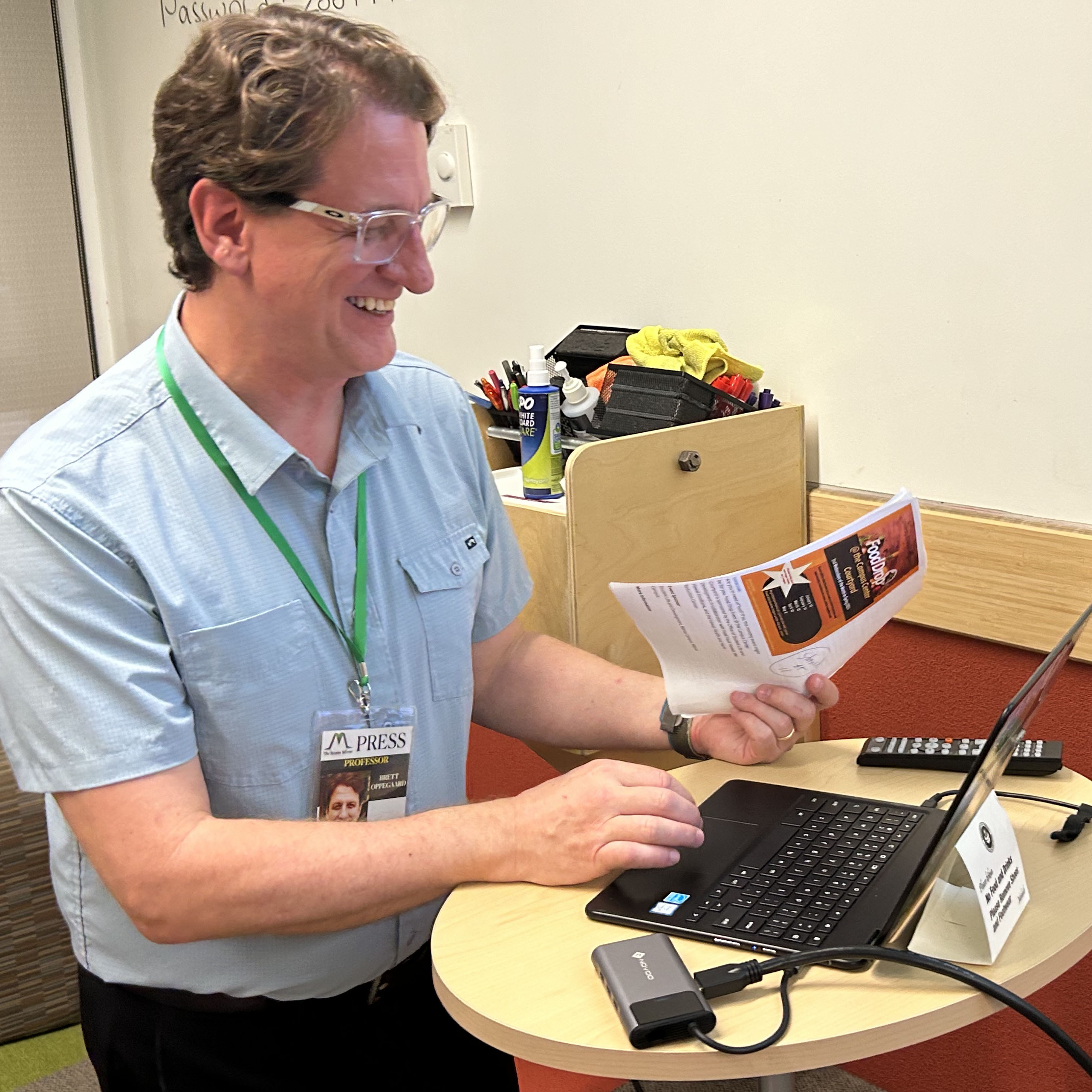 In this square color photograph, Brett Oppegaard, Principal Consultant at Access Hound, is shown in a profile portrait, looking at his computer and smiling. The 53-year-old man has short brown hair, blue eyes, and is wearing glasses with a clear frame. He is wearing a short-sleeved and light-blue collared shirt.