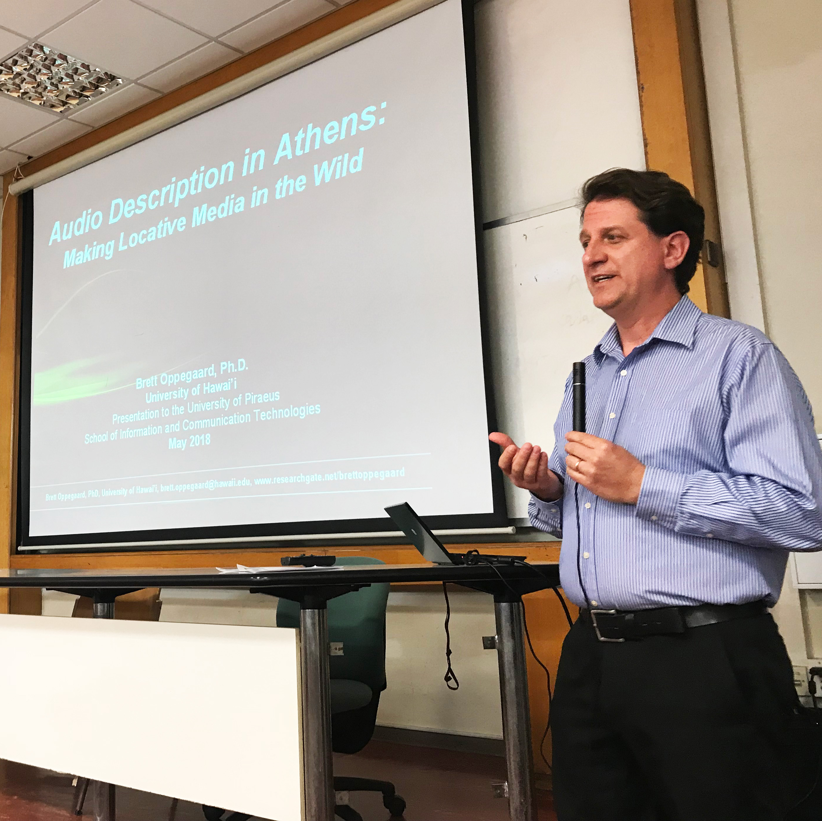 DESCRIBING: A square color photograph. Synopsis: Dr. Brett Oppegaard is standing at the front of a classroom, with a Powerpoint slide projected on the large screen behind him, titled 