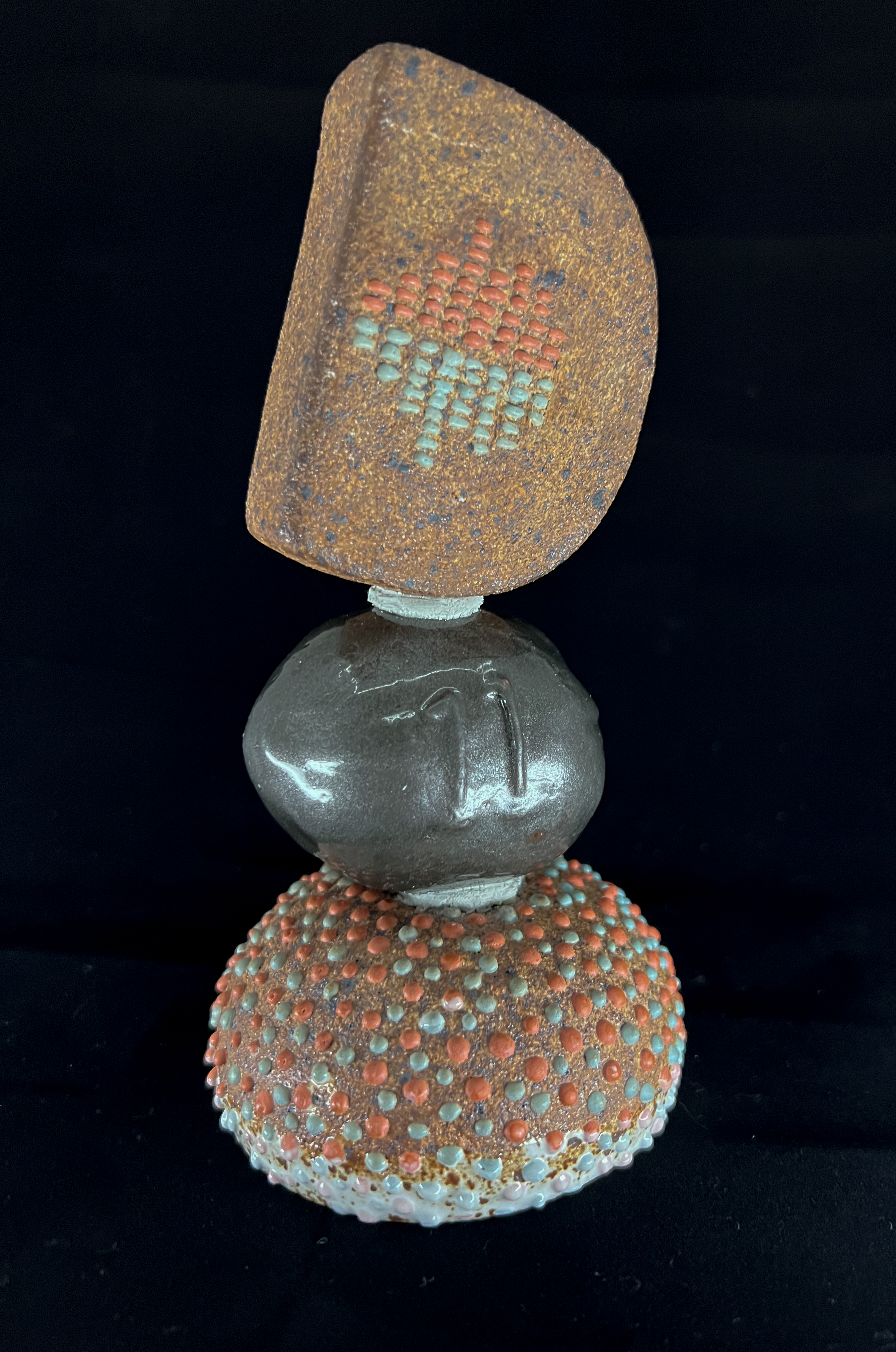 DESCRIBING: A vertical color photograph of the Descriptathon 11 Trophy. 
SYNOPSIS: A large upper-case D, The UniDescription Project's logo, rests slightly tilted to the right on top of a dark-gray coconut seed of sorts, which then rests on top of a pillow-shaped stone, reminiscent of a found object atop a sea urchin. All of the surfaces are heavily textured in rough material, including the counterspace in the D created by small horizontal rectangles — in orange and blue — resembling the sound waves on a digital stereo interface. The sea urchin pillow is covered with rounded orange and blue dots that are atop the surface of the sea urchin and touchable. A foam of white surf covers the bottom of the urchin, like it was found in low tide.