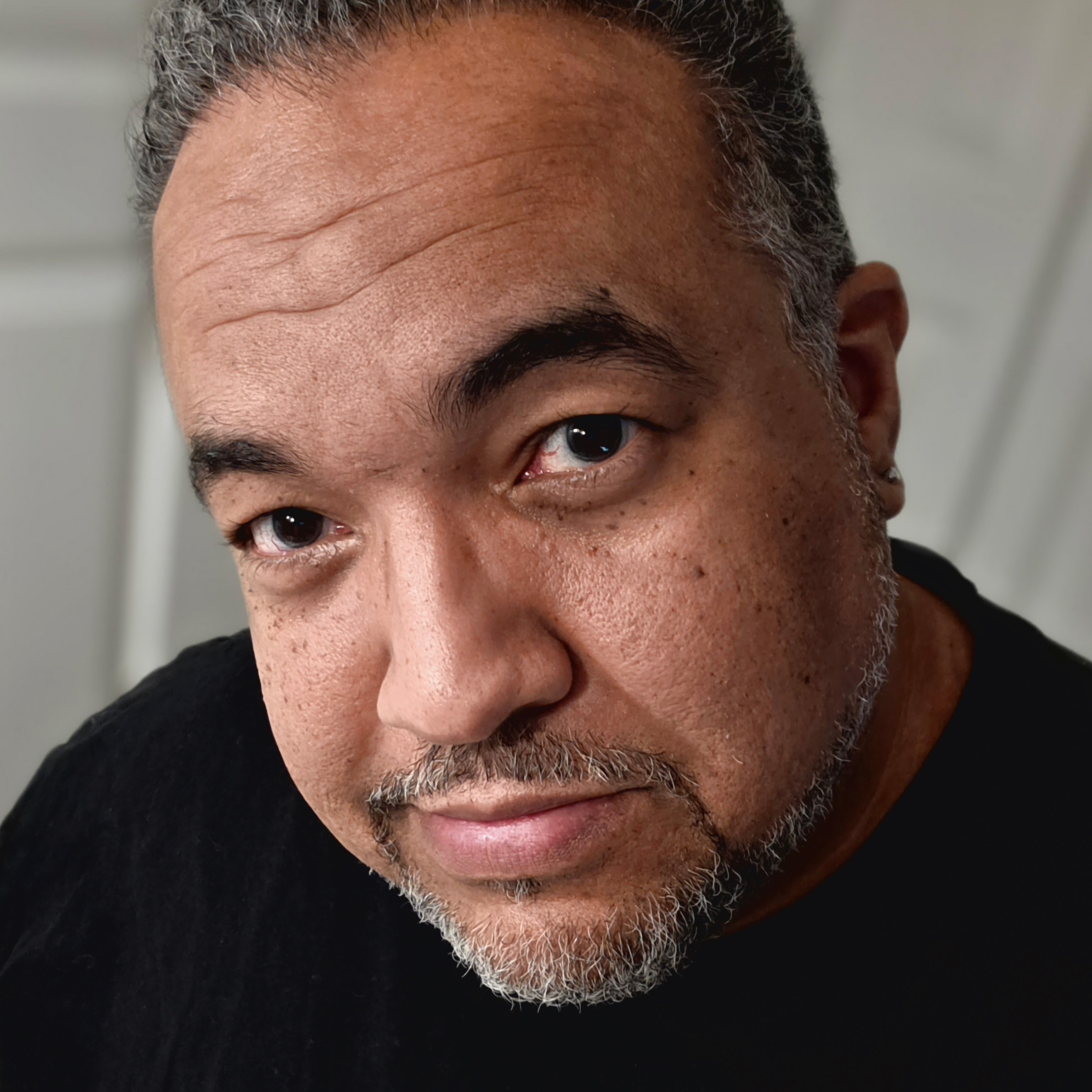 A light-skinned black man with curly salt and pepper shortcropped hair, beard and moustache, sits wearing a black crewneck sweater. He wears a slight smile, with his head slightly tilted to the side.