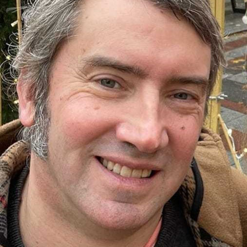 A Caucasian male of about 50 years old, with short brown hair that is graying. He is smiling and looking directly at the camera. He has blue eyes. The hood and plaid lining of a tan-colored coat is visible, as is the collar of an orange undershirt and a darker top on top of that.