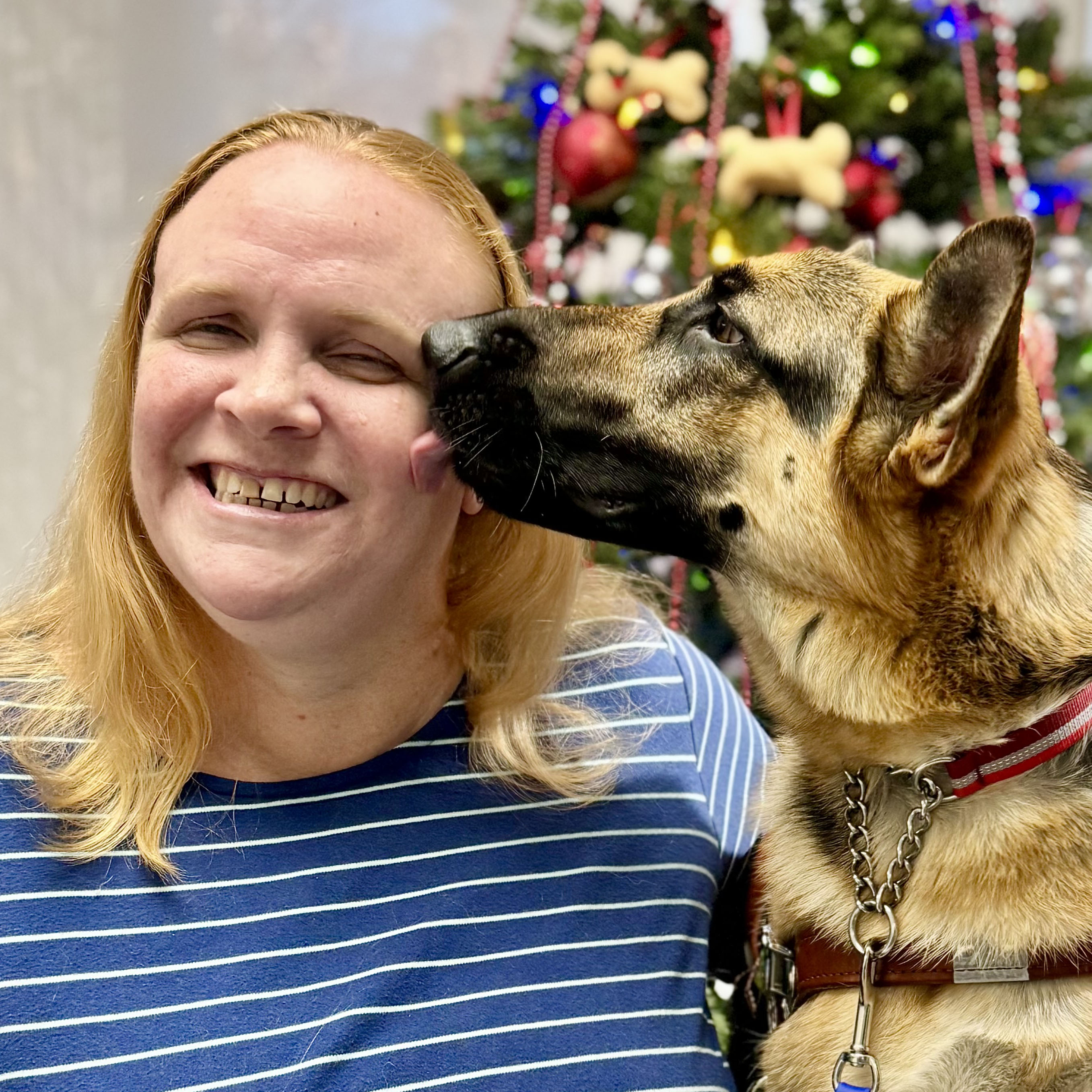 A photo of a woman with long strawberry blonde hair, wearing a long sleeve dark top with thin white stripes. Sitting next to her is a black and tan German Shepherd guide dog in harness. Behind them is a lit and decorated Christmas tree.
