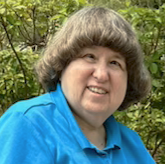 A vertical photo shows me, a 67 year old woman who is hard of hearing and blind. I'm smiling and sitting on a bench wearing a teal blue polo shirt. I have medium length curly brown hair.