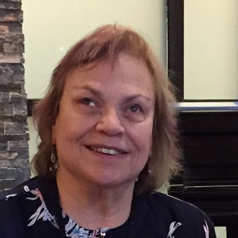 Caucasian woman of average height, hazel eyes, brown hair with blonde streaks wearing a shirt and decorated jeans with bright pink athletic shoes.