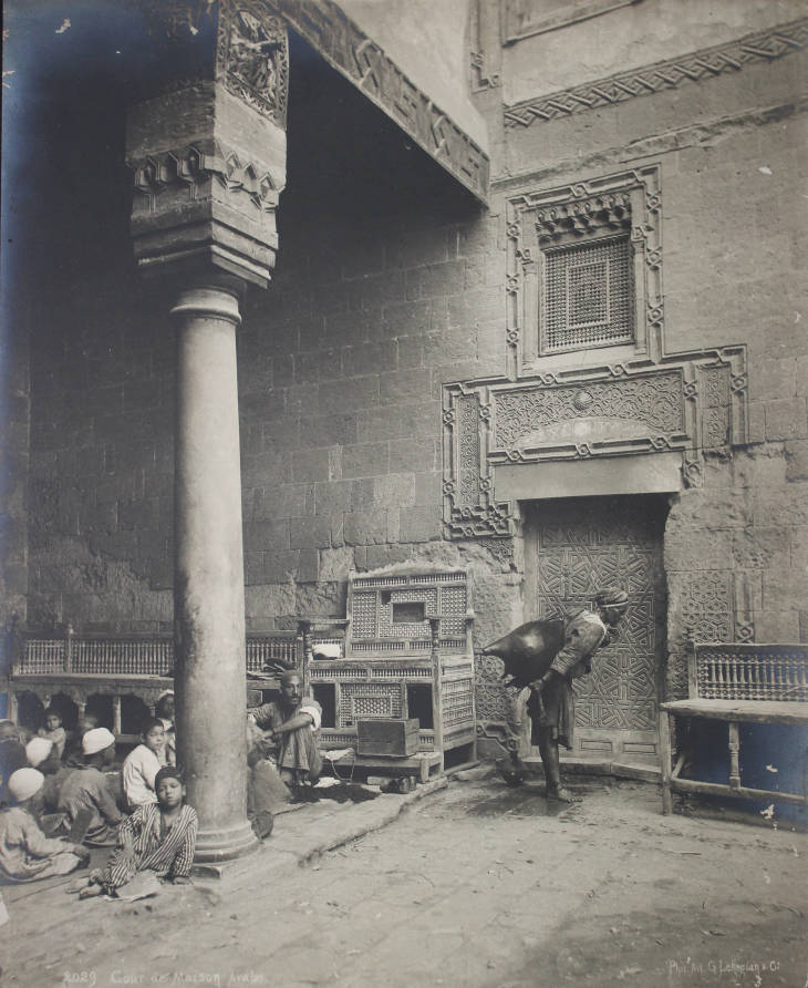 Title (English): A water carrier (Saqqa) in the courtyard of an Arab house. Title (Arabic): حاملة مياه (السقا) في فناء منزل عربي. Site (English): Saqqa Arab House Maison arabe. Site (Arabic): بيت السقا العربي. Collector: Creswell, K. A. C. (Keppel Archibald Cameron), 1879-1974. Date Created: ca. late 19th century or early 20th century.
