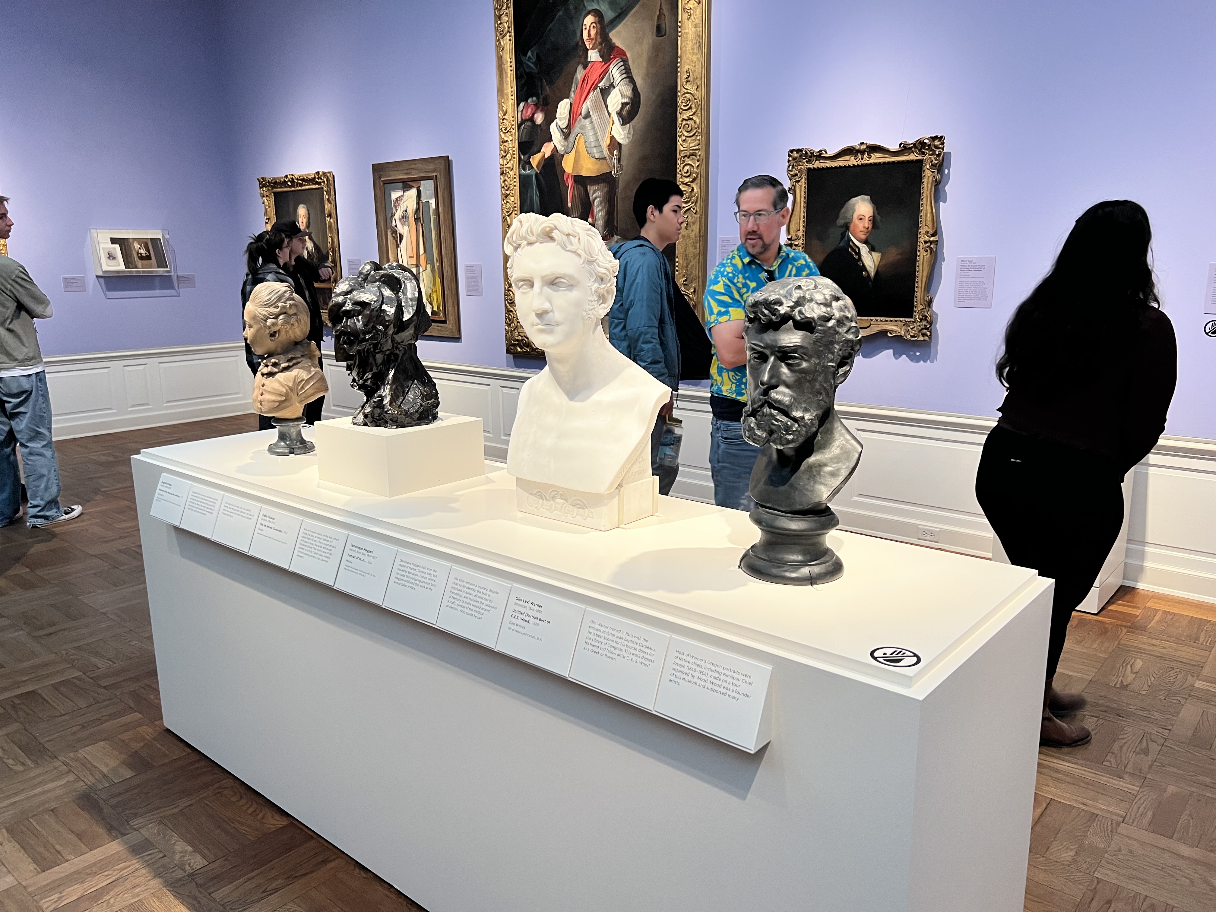 Four sculptural busts on display at the Portland Art Museum in Portland, OR, not boxed in by plexiglass. 

Describing: A horizontal color photograph.

Synopsis: A long white display pedestal in a gallery setting is topped with four sculptural busts, showing head-and-shoulder representations of four different people in different styles with different materials, from white marble to nearly black bronze. The second one to the viewer's left is Pablo Picasso's dark bronze "Tête de femme (Fernande)," recognizable at this distance by its fractional but interconnected pieces, creating the whole of the head. The labels for the other busts cannot be read in this image from this distance. 

IN-DEPTH DESCRIPTION: The white pedestal crosses the frame of the photo diagonally, from about mid-frame on the left side to nearly the bottom right corner, so the bust on the far left appears smaller, and the one of the right appears larger. The front edge of the pedestal has a long rectangular strip of signage at the top, labeling and describing each piece of artwork. Near the front-right corner of the pedestal, a small round sign on the top indicates a gallery rule: No touching.
On the pedestal, four busts are spaced roughly a foot apart. From the viewer's left to right, 1. A small beige bust of a person with curled hair and a high-collared, buttoned coat on a short gray stand; 2. A dark, rough-textured, modern-looking bust with angular planes and ridges (the Picasso); 3. A white, smooth bust, larger than the others, with short wavy hair and neutral expression, set on a square base; and, 4. A dark metal bust of a bearded man with a furrowed brow on a rounded plinth.
Behind them, the gallery's walls are violet, and several framed paintings are displayed, including, at center, a tall, ornate, gold-framed portrait of a standing figure wearing a red sash; to its right, a smaller, gold-framed portrait of a man shown from the shoulders up, with gray hair and a military jacket; to its left, smaller works also are present, including at least one with geometric shapes rather than traditional naturalistic portraiture. A half-dozen visitors in casual clothes stand near the wall and pedestal, reading labels or chatting.