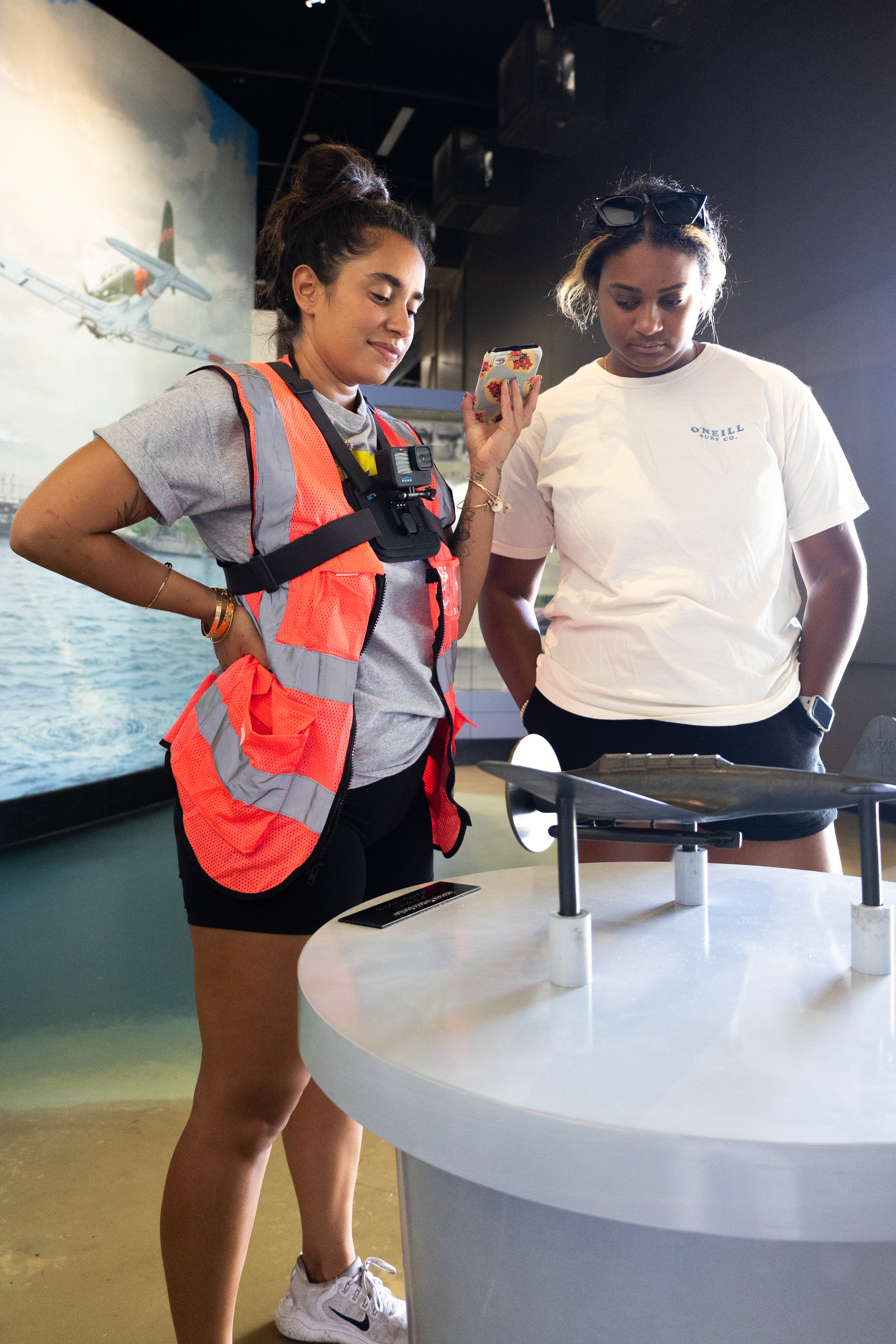 Two brown-skinned young women stand near a small bronze sculpture of a Japanese Zero plane on a pedestal, with a painted version of the same plane on the wall behind them. The woman on the left, in an orange safety vest with a camera strapped to her chest and long black hair in a high bun, holds an audio-recording device toward the second woman. The woman on the right, with sunglasses pushed up on her head and dark hair in a ponytail, listens intently to the audio description playing about the plane.