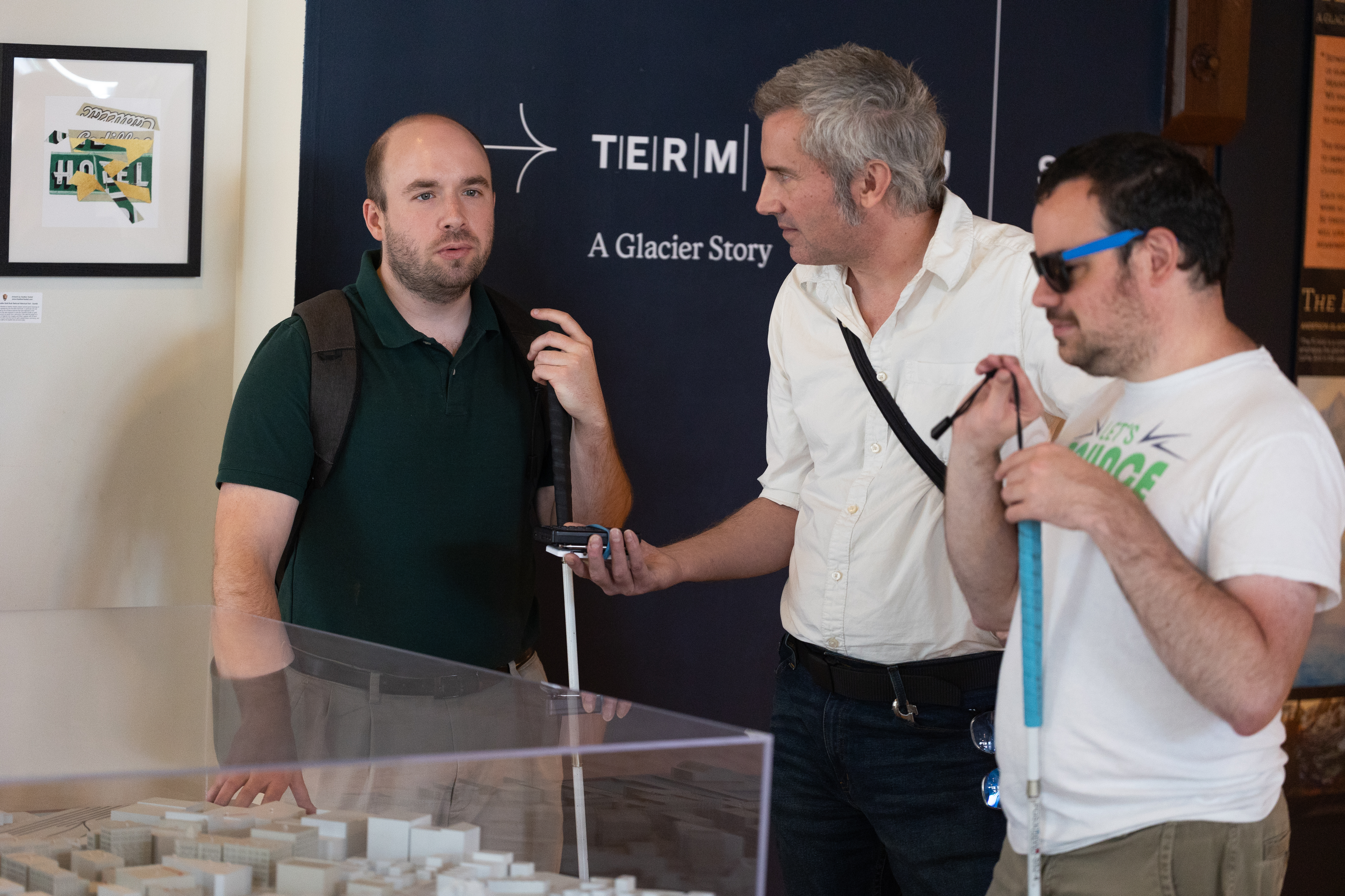 Describing: A horizontal color photograph showing three white men standing near a museum display. Description: Access Hound's Matt Bullen, center, with short gray hair, stands talking with two young men, one to his left and one to his right, both holding white canes, indicating that they are blind. Bullen holds an audio recorder to capture their comments as he talks with them about their museum surroundings. Behind the group is a wall with a partially obscured display that reads 'A Glacier Story,' with another wall decoration saying, 'The Cadillac Hotel.'