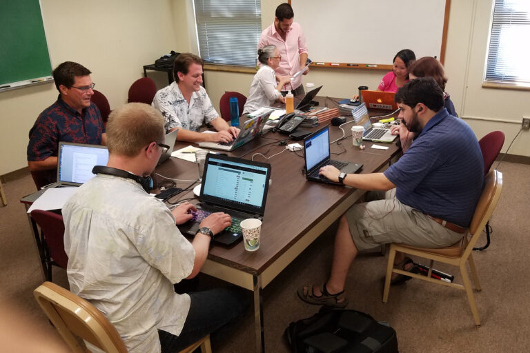 DESCRIBING: A horizontal color photograph. DESCRIPTION: The second virtual Descriptathon had a large support team working behind the scenes on it. This image shows a conference room in Crawford Hall at the University of Hawai'i, where most of the core group of Descriptathon administrators gathered to host the 28-team event. These eight people are sitting around a rectangular table. If you imagine the table as sort of a Daliesque clock, with Research Assistant Tuyet Hayes, in a bright pink shirt at the Noon position, the rest of the team, clockwise, is: Consultant Annie Leist, Consultant Sina Bahram, Developer Joe Oppegaard, Co-PI Thomas Conway, PI Brett Oppegaard, NPS Accessibility Coordinator Michele Hartley, and Research Assistant Phil Jordan. Everyone has a laptop in front of them and is busily working, except Jordan, who is standing next to Hartley and talking with her about a piece of paper he is holding.