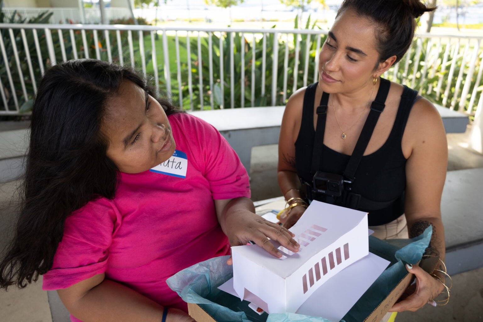 Volunteer participant Crata Mizutani, left, studies the shape of the USS Arizona Memorial building by touching a paper model of it provided by UniD researchers, including Research Assistant Kira Swearingen. Swearingen watches Mizutani examine the model while Mizutani, who is blind, provides think-aloud reactions to what sheʻs feeling and what sheʻs already learned that November 2023 day about the structure through Audio Description. This photograph, which has a horizontal orientation, shows Mizutani seated and leaning to her right while feeling the model in her lap with her left hand. Sheʻs wearing a bright pink t-shirt, and she has long, black hair. Swearingen, wearing a Go Pro camera around her neck to record Mizutaniʻs actions and observations, is seated next to Mizutani, on her left. Swearingen is looking carefully and kindly at Mizutani, studying her movements and listening to her comments. Swearingen is wearing a black tank top, and she has her long hair in a bun.