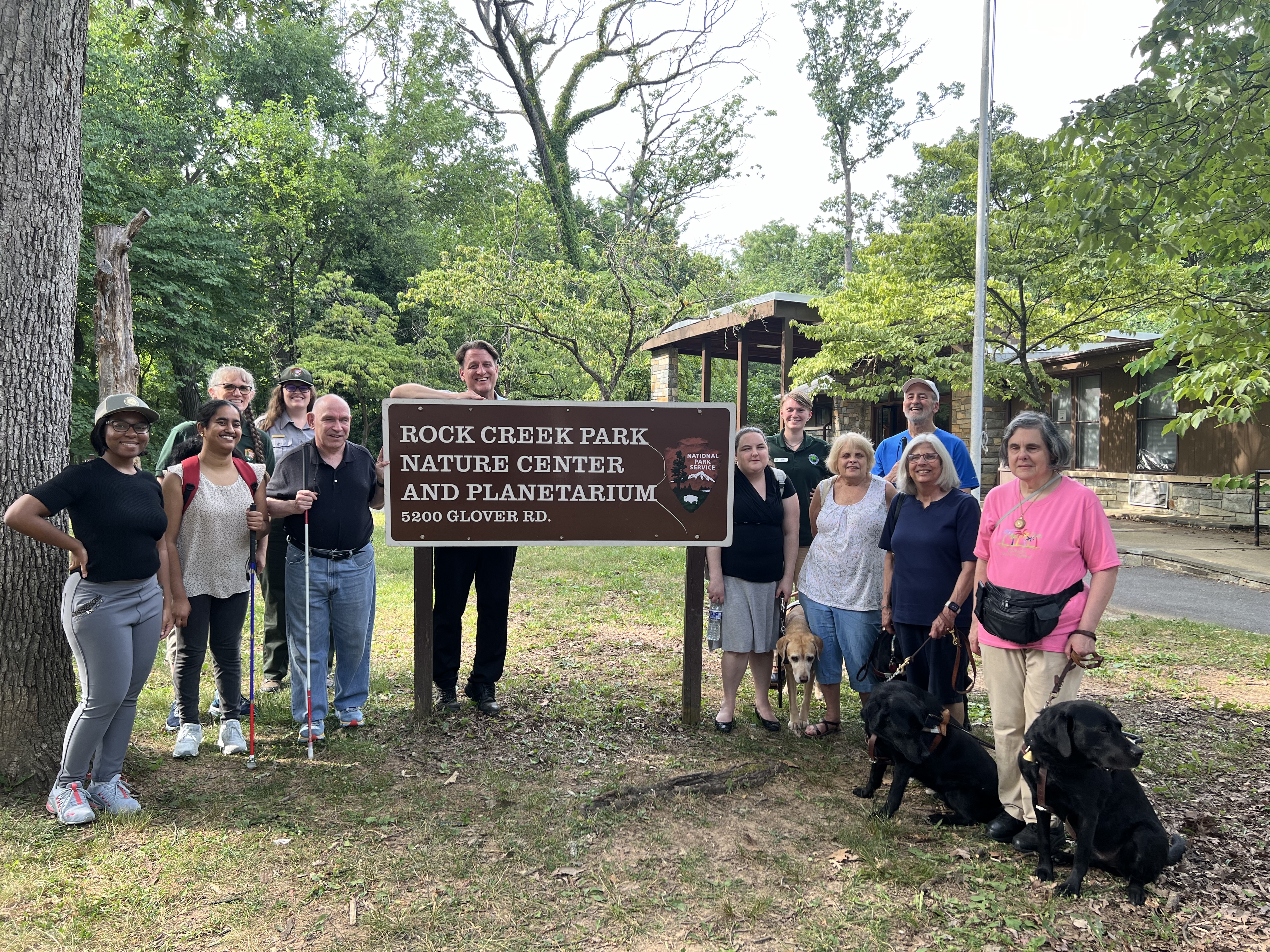 DESCRIBING: A horizontal color photograph taken June 25, 2024. DESCRIPTION: A group photo of a dozen of the participants in a UniDescription field-research test at Rock Creek Park, standing around a large brown and metal sign that reads 'Rock Creek Park Nature Center and Planetarium, 5200 Glover RD.' In the background of this scene, there is a wooded area, filled with trees and a large building that serves as a visitor center. Directly behind this metal sign is Dr. Brett Oppegaard, the principal investigator of the UniDescription project and a professor from the University of Hawaii. He is a 53-year-old white man with short brown hair. His head can be seen above the sign, and his legs, in black pants and with black shoes can be seen below the sign. He facilitated the discussions during the event. Behind him, and to his right, are two NPS staff members who also were involved in organizing the field trip: Autumn Cook, a U.S. National Park Service Public Affairs Specialist at Rock Creek Park, and Tera Lynn Gray, an Accessibility Specialist at the Harpers Ferry Interpretive Design Center. American Council of the Blind members pictured are: Swatha Nandhakumar, ACB's Advocacy and Outreach Specialist, Claire Stanley, ACB's Director of Advocacy and Governmental Affairs, and members Pat Sheehan, Cindy LaBon, Susan Crawford, Doug Powell, and Jane Carona, along with three guide dogs. In the photo as well are NPS interns Cameron Waiters and Jordan Land, who helped with the on-site logistics of the event.