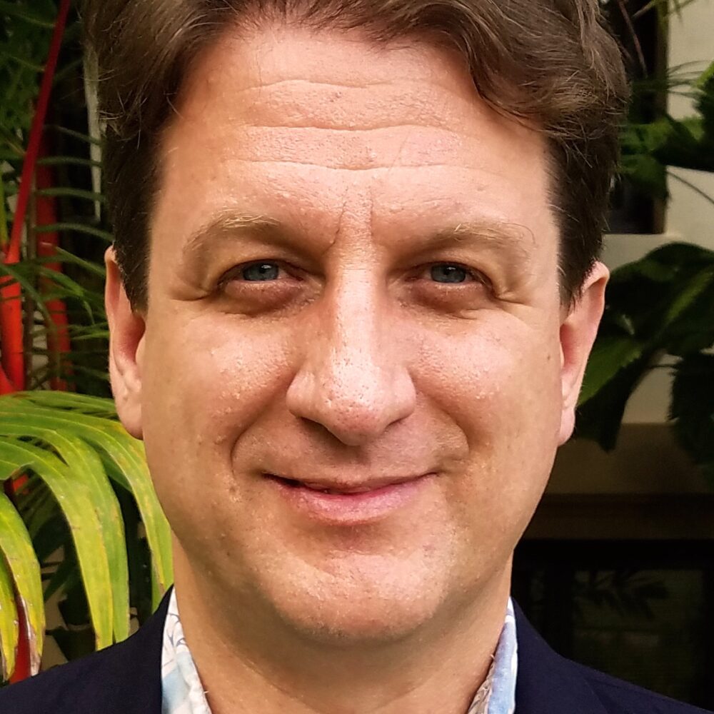 In this small vertical portrait, I am shown from my shoulders up to roughly the middle of my forehead. I am a 53-year-old American man, white in the abstract but with ancestral origins in the United Kingdom and Norway. Indicating those roots visually, I have blue eyes, a pinkish-tan skin with a red undertone, and dark brown hair. My hair is short on the sides but kept long on the top, showing some waviness to it. I do not have any facial hair beyond my eyebrows. Contextually, I am 6 feet tall and weigh about 210 pounds, but there is nothing in this image to use to gauge my body’s size or shape. The collar of my light-blue and patterned Aloha shirt shows just above my dark-blue suit jacket. On the UH campus, Aloha shirts are common, and almost no one wears a tie. So I’m dressed for that work environment. I am looking directly into the camera and expressing a subtle smile, with my lips slightly upturned at the corners. I am hoping to convey warmth and friendliness while still maintaining an authentic facial expression.